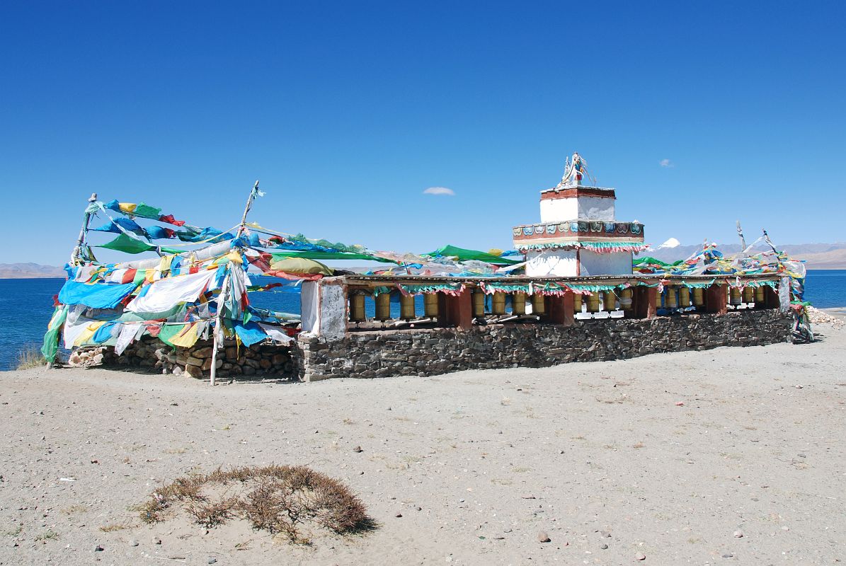 16 Seralung Gompa Chorten And Prayer Wheels With Lake Manasarovar And Mount Kailash Beyond Seralung Gompa has a chorten next to Lake Manasarovar with a row of prayer wheels and Mount Kailash in the distance.